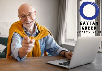 Fashionable friendly elderly male recruiter wearing rectangular glasses and elegant clothes working on portable computer, smiling broadly and pointing finger at camera, choosing you for job position